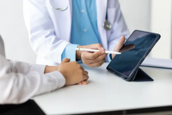 Doctor in white coat using a stylus to explain information on a tablet to a patient at a white desk.