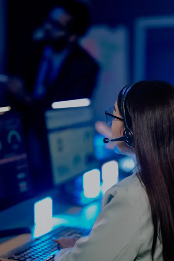 Woman wearing a headset looking at multiple computer monitors in a blue-lit control room