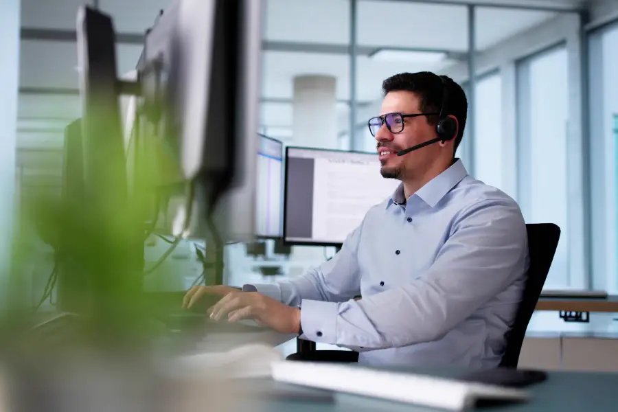 Smiling man wearing a headset and glasses working on a computer in a bright office