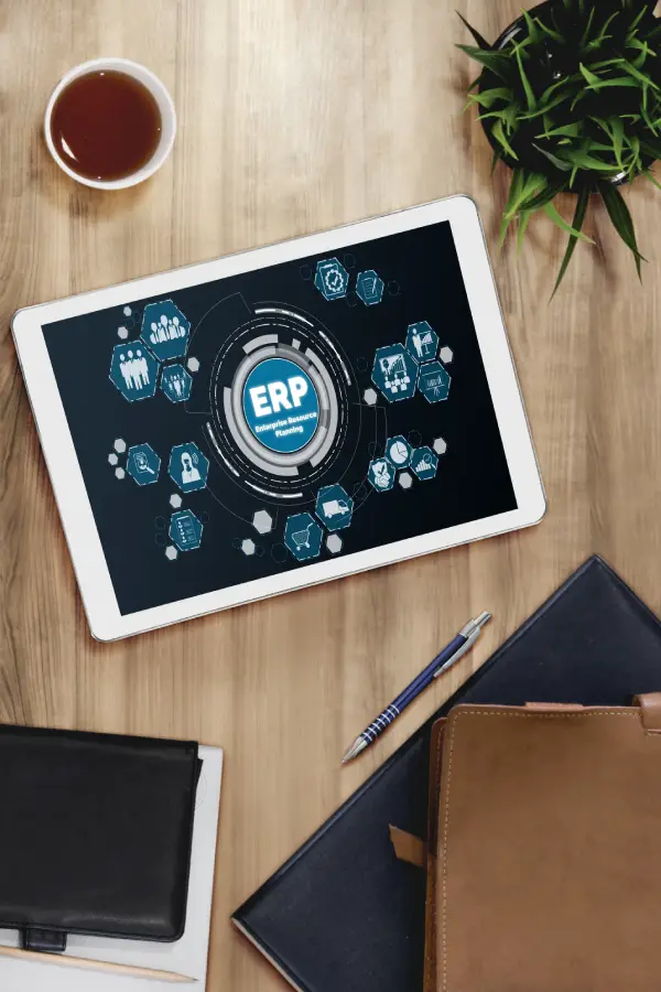 Top-down view of a tablet displaying an ERP system interface on a wooden desk with a coffee cup, notebooks, and a pen