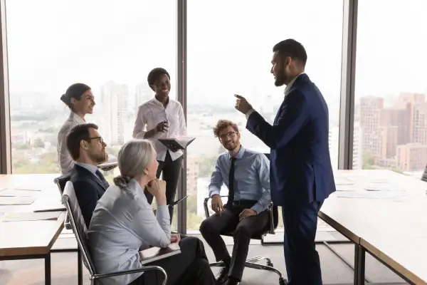 Businessman in a blue suit speaking to seated colleagues during a meeting in a sunlit conference room