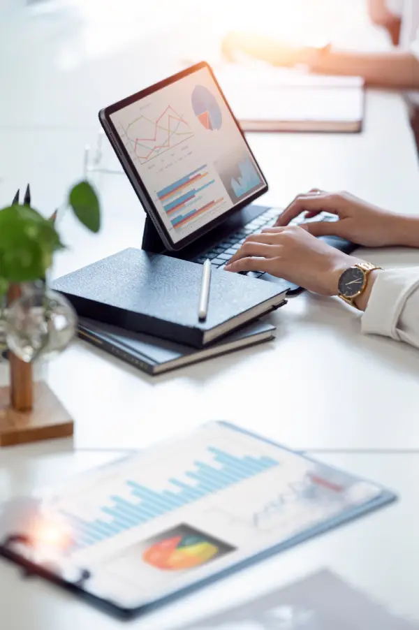 A person typing on a tablet keyboard with charts on screen, notebooks with pen nearby, and documents with graphs on a white desk.