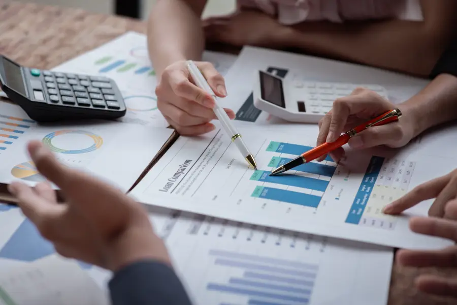 Hands of three people analyzing financial charts and graphs with pens and calculators on a desk.