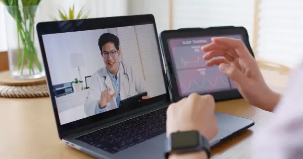 A person using a laptop for a telehealth consultation with a healthcare professional visible on the screen