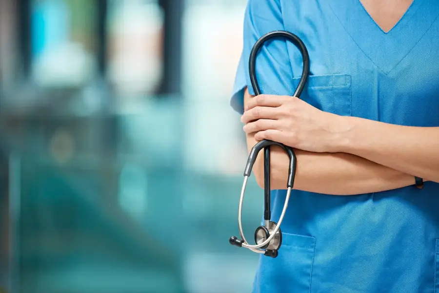 Healthcare worker in blue scrubs holding a stethoscope with arms crossed against a blurred clinical background.