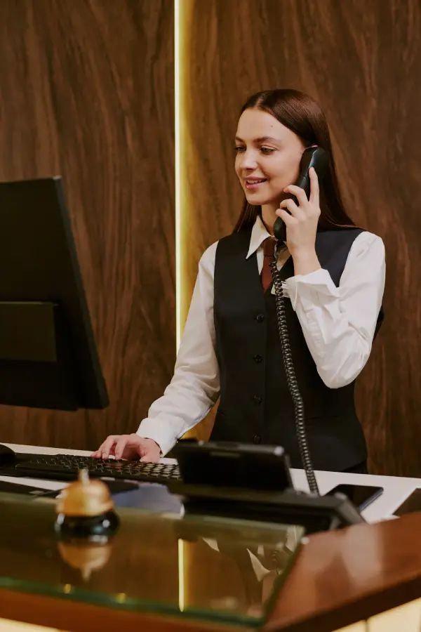 Receptionist in a black vest speaking on the phone while using a computer at a hotel front desk with a ringing bell on the counter.