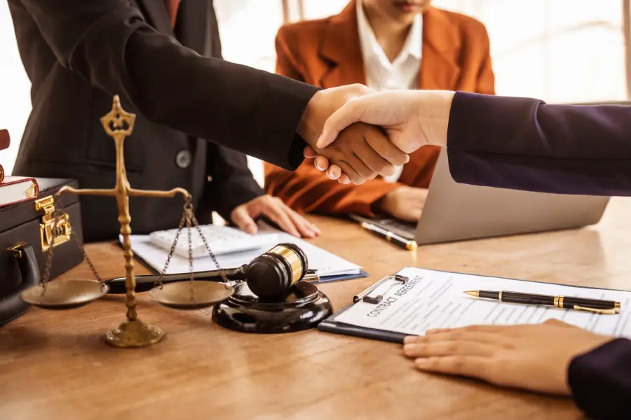 Professionals shaking hands across a desk with a scale of justice, a gavel, and contract documents