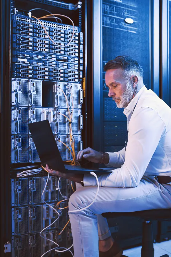 IT professional sitting on a chair using a laptop while connected to a large server rack in a blue-lit server room