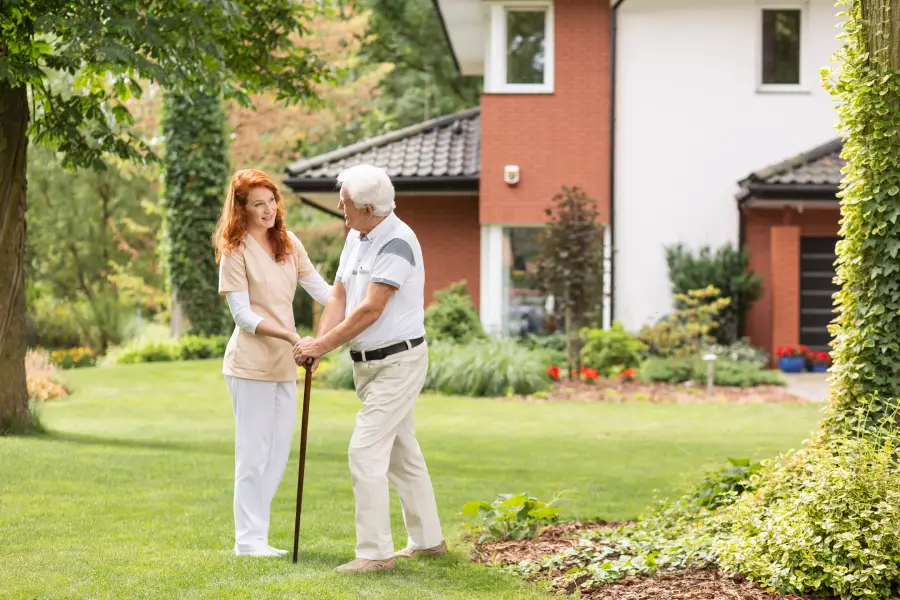Caregiver assisting an elderly man walking with a cane outside a brick house