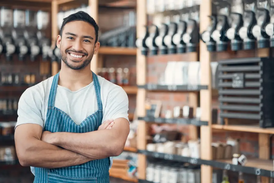 Smiling male barista with crossed arms wearing a blue striped apron in a coffee shop