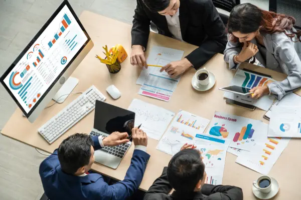 Four business professionals reviewing charts and graphs on paper and digital devices during a meeting at a desk.