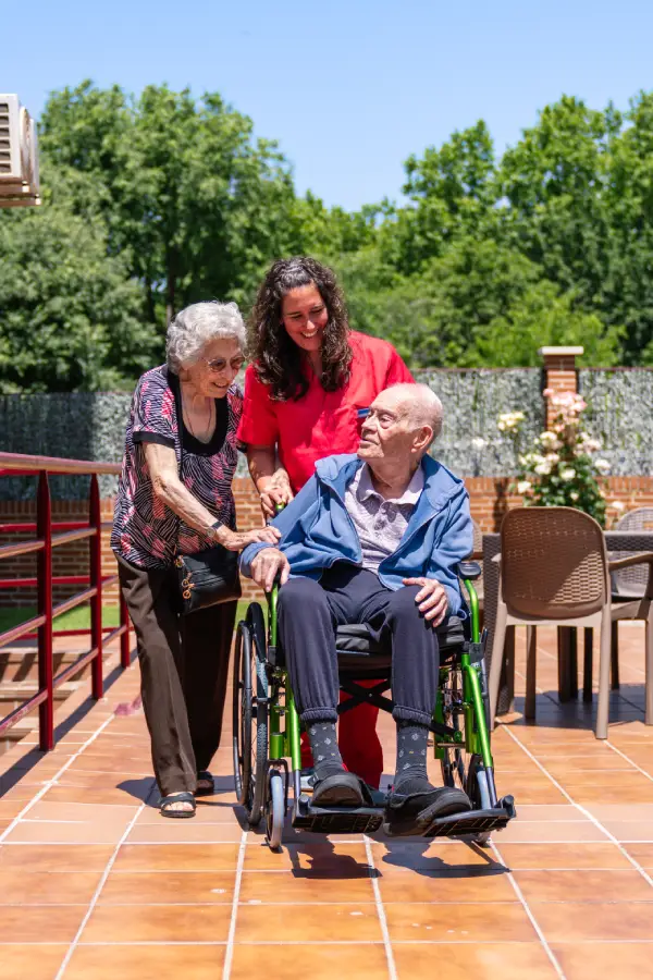 Caregiver pushing an elderly man in a wheelchair on a sunny patio, with an older woman walking beside them