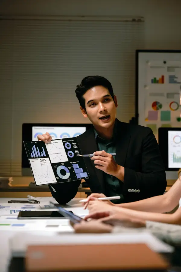 Man in a business meeting presenting charts and data on a tablet to colleagues seated at a conference table.